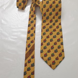 May include: Mustard yellow tie with a repeating pattern of small, colorful snail illustrations. The tie features a brown label with the brand name "Longchamp". The tie is made of a smooth fabric.
