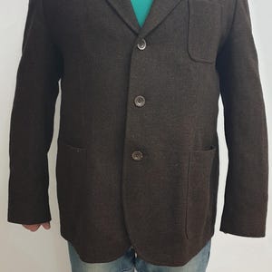 Puede incluir: Una chaqueta de tweed marrón con cierre de tres botones y dos bolsillos de solapa. La chaqueta tiene un frente liso y una tela ligeramente texturizada.