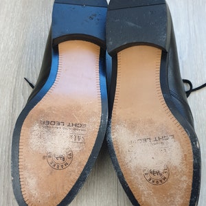 May include: Close-up of the soles of two black leather shoes. The soles are brown and have the text "MARKE" and "ECHT LEDER" printed on them.