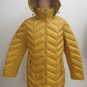 May include: Mustard yellow puffer jacket with a hood. The jacket features a full-length zip, two side pockets, and a quilted pattern. The jacket is displayed on a mannequin. A winter outerwear item.