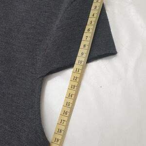 Puede incluir: Un su&eacute;ter gris oscuro de manga corta con una cinta m&eacute;trica al lado. La cinta mide la longitud de la manga, mostrando aproximadamente 23 cm. El su&eacute;ter tiene cuello redondo y parece estar hecho de tejido de punto.