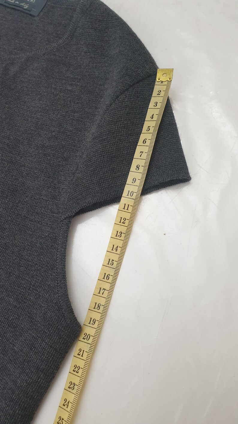 Puede incluir: Un su&eacute;ter gris oscuro de manga corta con una cinta m&eacute;trica al lado. La cinta mide la longitud de la manga, mostrando aproximadamente 23 cm. El su&eacute;ter tiene cuello redondo y parece estar hecho de tejido de punto.