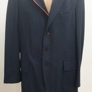 Könnte beinhalten: Ein marineblauer Blazer mit fallendem Revers, Dreiknopfverschluss und zwei Vordertaschen. Der Blazer hat einen roten Reißverschluss im Innenfutter. Die Jacke wird auf einer Schaufensterpuppe präsentiert.