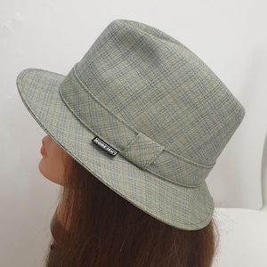 Puede incluir: Un sombrero fedora a cuadros grises con ala estructurada y una banda alrededor de la corona. El sombrero tiene una pequeña etiqueta negra que dice "GORE-TEX". El sombrero está sobre una cabeza de maniquí.
