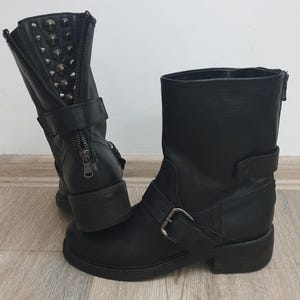 Könnte beinhalten: Ein Paar schwarze Leder-Ankle-Boots. Einer der Stiefel hat silberfarbene Nieten und einen Reißverschluss. Der andere Stiefel hat eine Schnalle und einen kleinen Absatz. Die Stiefel stehen auf einer hellen Holzmaserung.