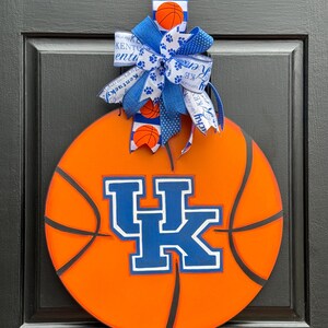 May include: Orange basketball-themed door hanger featuring the University of Kentucky Wildcats logo in blue and white. The door hanger is adorned with a blue and white bow with basketball and paw print accents. The door hanger is hanging on a black door.