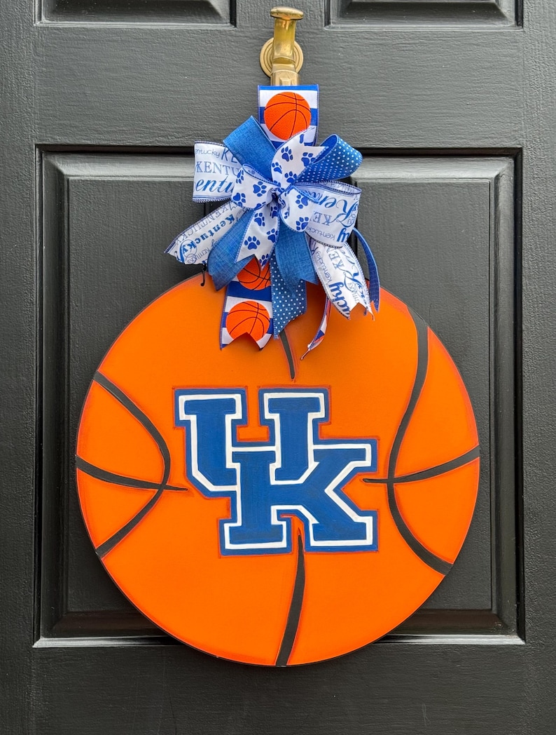May include: Orange basketball-themed door hanger featuring the University of Kentucky Wildcats logo in blue and white. The door hanger is adorned with a blue and white bow with basketball and paw print accents. The door hanger is hanging on a black door.
