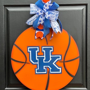 May include: Orange basketball-shaped door hanger with black lines and the letters "UK" in blue and white. A blue and white bow with basketball and paw print accents is attached to the top. The door hanger is hanging on a black door.