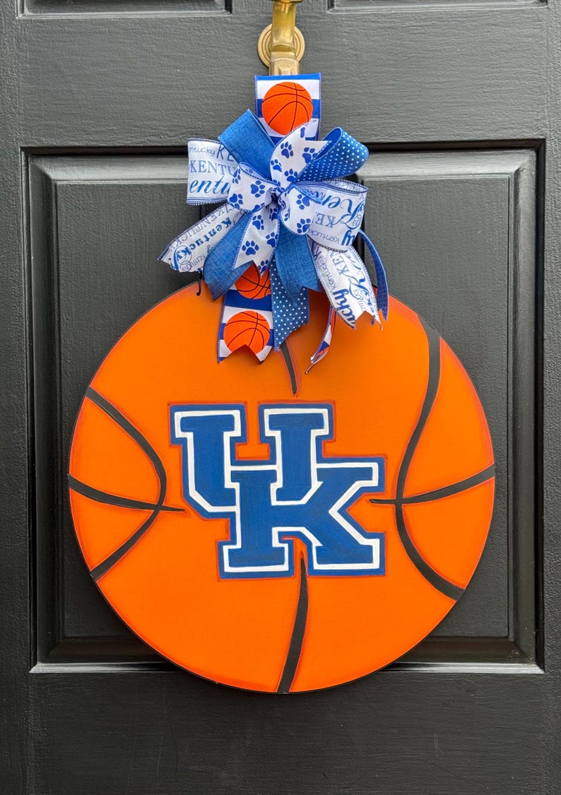 May include: Orange basketball-shaped door hanger with black lines and the letters "UK" in blue and white. A blue and white bow with basketball and paw print accents is attached to the top. The door hanger is hanging on a black door.