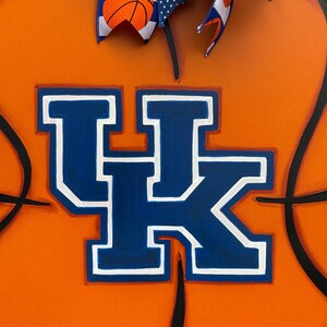May include: A wooden basketball-themed door hanger. The orange background features black lines mimicking a basketball. The center displays the blue and white letters "UK". A blue and white ribbon with basketball and polka dot patterns adorns the top.