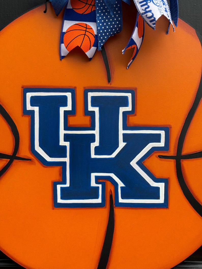 May include: A wooden basketball-themed door hanger. The orange background features black lines mimicking a basketball. The center displays the blue and white letters "UK". A blue and white ribbon with basketball and polka dot patterns adorns the top.