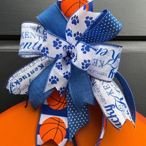 May include: A decorative bow featuring blue and white ribbons with basketballs and paw prints. The bow has a Kentucky theme, with text that reads "Kentucky". The bow is attached to an orange surface.