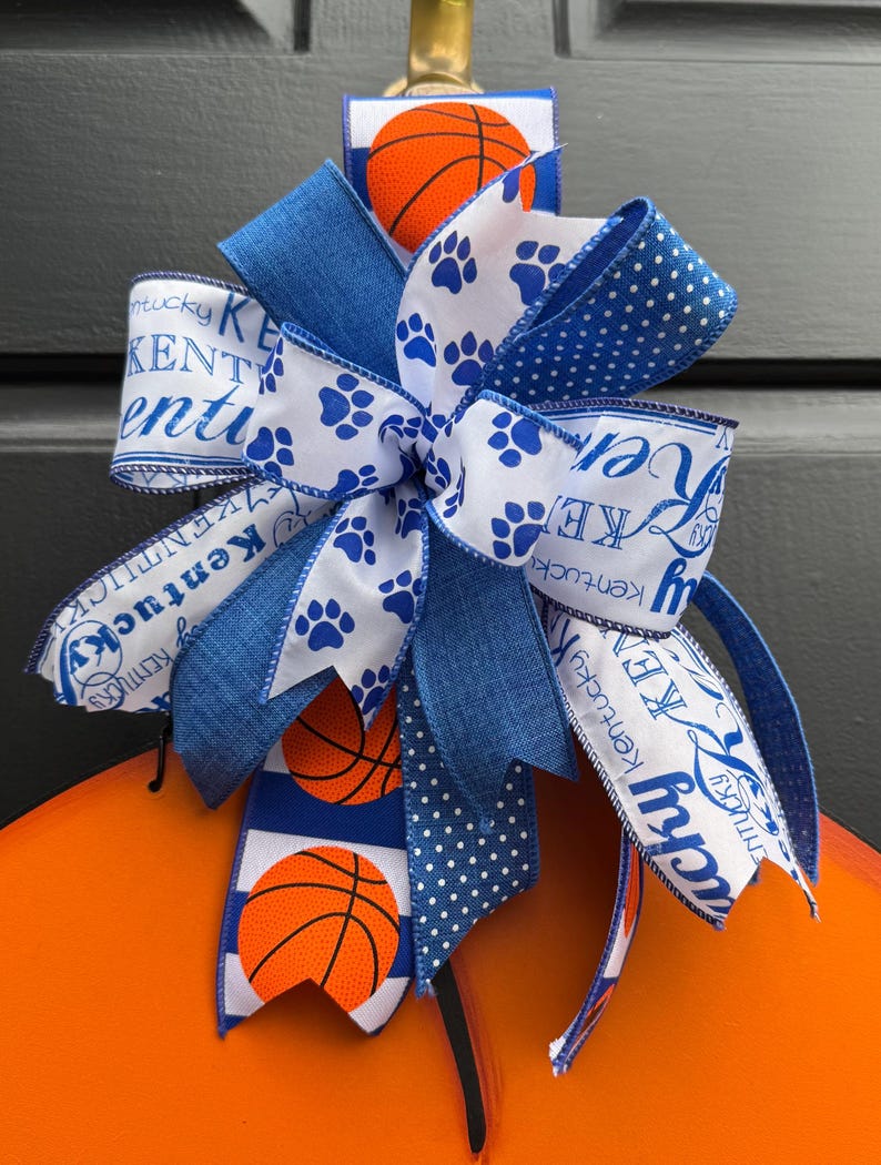 May include: A decorative bow featuring blue and white ribbons with basketballs and paw prints. The bow has a Kentucky theme, with text that reads "Kentucky". The bow is attached to an orange surface.