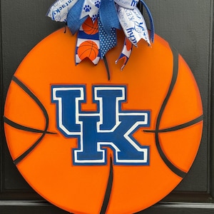 May include: Round orange basketball-themed door hanger with black lines and the blue and white "UK" logo. A blue and white bow with basketball and paw print accents tops the door decoration.