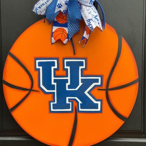 May include: Round orange basketball-themed door hanger with black lines and the blue and white "UK" logo. A blue and white bow with basketball and paw print accents tops the door decoration. A decorative item for sports fans.