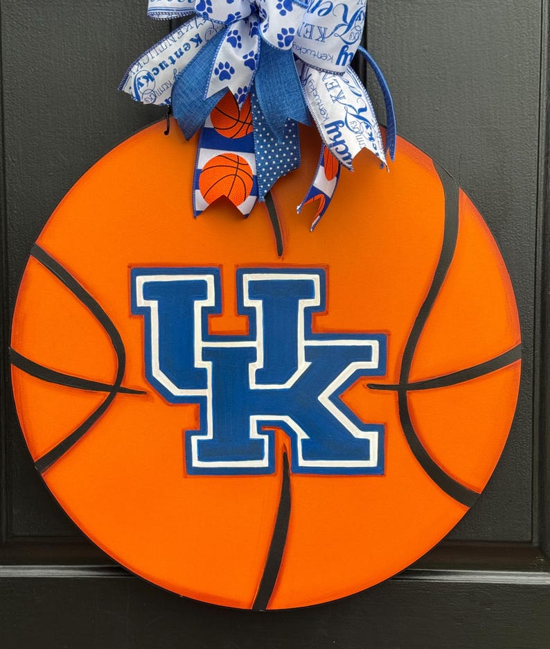May include: Round orange basketball-themed door hanger with black lines and the blue and white "UK" logo. A blue and white bow with basketball and paw print accents tops the door decoration. A decorative item for sports fans.
