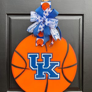May include: A round, orange basketball-themed door hanger featuring the University of Kentucky Wildcats logo in blue and white. The design includes black lines mimicking a basketball and a decorative bow.