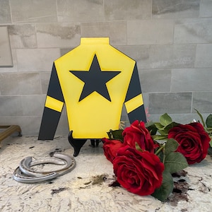 May include: A yellow and black jockey silks with a black five-pointed star in the center. The silks are displayed on a wooden stand with three red roses in the foreground.