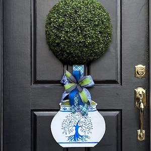 May include: A blue and white ceramic planter with a tree design and a green and blue bow. The planter is hanging on a black door with a green topiary ball above it.