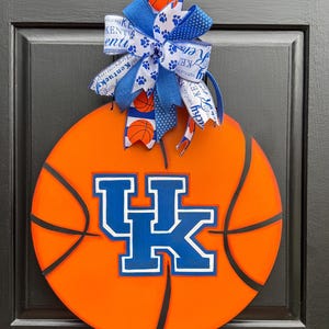 May include: A basketball-themed door decoration featuring an orange basketball design with black lines. The center displays a blue and white "UK" logo. A blue and white bow with paw prints and basketball accents tops the decoration.