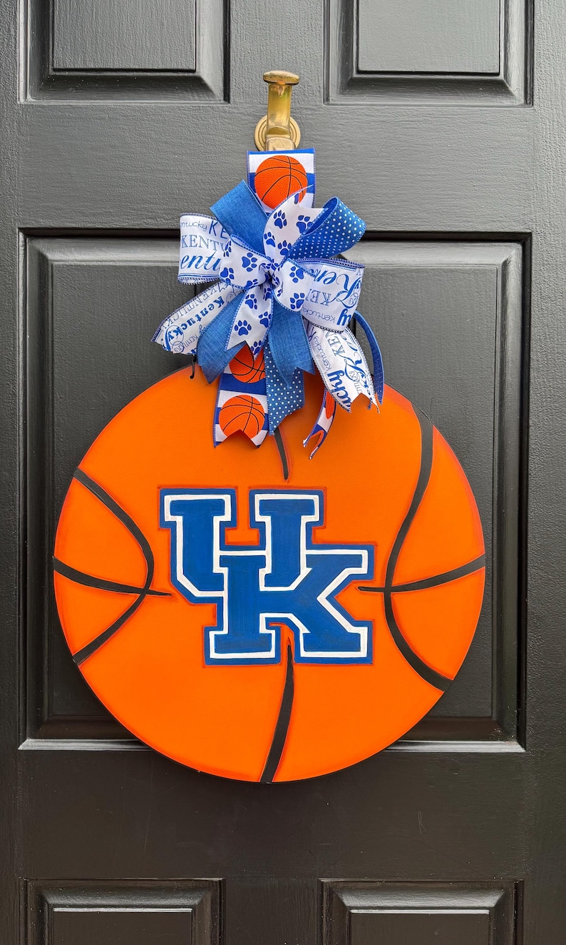 May include: A basketball-themed door decoration featuring an orange basketball design with black lines. The center displays a blue and white "UK" logo. A blue and white bow with paw prints and basketball accents tops the decoration.