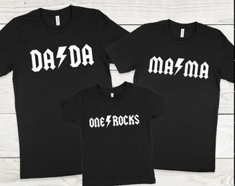May include: Three black t-shirts with white text. The largest shirt says "DA DA" with a lightning bolt between the words. The medium shirt says "MA MA" with a lightning bolt between the words. The smallest shirt says "ONE ROCKS" with a lightning bolt between the words.