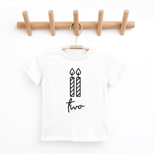 May include: White t-shirt with a black graphic of two birthday candles and the word "two" written in cursive.
