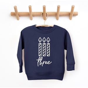 May include: Navy blue sweatshirt with a white graphic of three candles and the word "three" in a script font. The sweatshirt is hanging on a wooden coat rack against a white background.