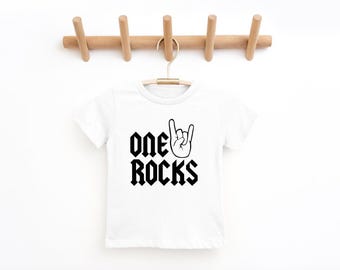 May include: White t-shirt with the text "ONE ROCKS" in a bold, black gothic font, accompanied by a hand gesture. The shirt is hanging on a wooden hanger against a white background. A fun and stylish tee for kids.