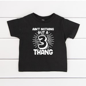 May include: Black toddler t-shirt with white text that reads "Ain't Nothing But a 3 Thang." The number 3 is in the center of the design, surrounded by a burst of lines. The shirt is displayed on a white wooden surface.