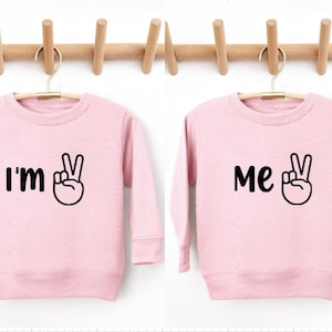 May include: Two pink long-sleeved shirts with black text and a peace sign graphic. The shirt on the left says "I'm" and the shirt on the right says "Me".