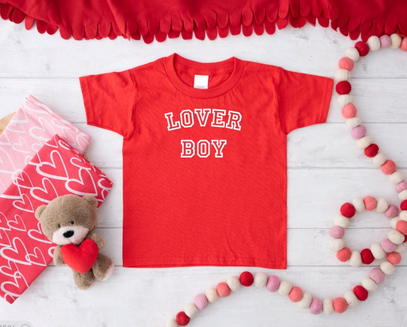 Puede incluir: Camiseta roja con texto blanco que dice "LOVER BOY" en una fuente de estilo universitario.
