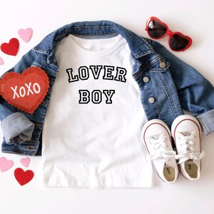 Puede incluir: Camiseta blanca con texto negro que dice "LOVER BOY" en una fuente de estilo universitario. La camiseta est&aacute; colocada sobre una superficie blanca con una chaqueta vaquera, gafas de sol rojas en forma de coraz&oacute;n y decoraciones rojas y rosas en forma de coraz&oacute;n.