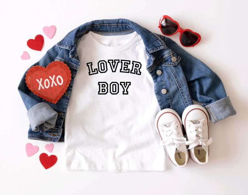 Puede incluir: Camiseta blanca con texto negro que dice "LOVER BOY" en una fuente de estilo universitario. La camiseta est&aacute; colocada sobre una superficie blanca con una chaqueta vaquera, gafas de sol rojas en forma de coraz&oacute;n y decoraciones rojas y rosas en forma de coraz&oacute;n.