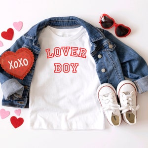 Puede incluir: Camiseta blanca con texto rojo que dice "LOVER BOY" en una fuente de estilo universitario. La camiseta est&aacute; colocada sobre una superficie blanca con una chaqueta vaquera, gafas de sol rojas en forma de coraz&oacute;n y corazones de fieltro rojo.