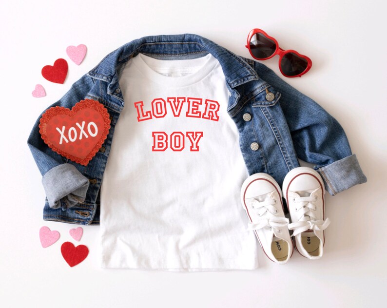 Puede incluir: Camiseta blanca con texto rojo que dice "LOVER BOY" en una fuente de estilo universitario. La camiseta est&aacute; colocada sobre una superficie blanca con una chaqueta vaquera, gafas de sol rojas en forma de coraz&oacute;n y corazones de fieltro rojo.