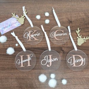 May include: Five clear round acrylic ornaments with white ribbon hangers. Each ornament has a different name written in white script with a gold initial. The names are:  'Hannah', 'Jason', 'Danny', 'Cameron', and 'Kerrie'.
