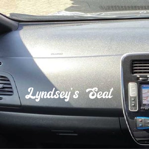 May include: A white vinyl sticker with the text " Lyndsey's Seat" on a gray car dashboard.