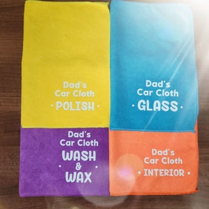 May include: Four microfiber cloths in yellow, blue, purple, and orange with white text. The text reads "Dad's Car Cloth" followed by a cleaning task: "POLISH", "GLASS", "WASH & WAX", and "INTERIOR".
