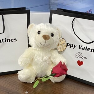 May include: Two white gift bags with black trim and handles. The bags have the text "Happy Valentines" printed on them, with a name and a red heart below. A white teddy bear with a red rose is sitting in front of the bags.
