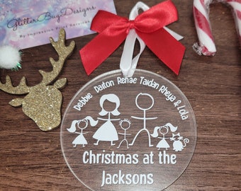 May include: A clear, circular Christmas ornament with a red bow and white ribbon. The ornament features a family stick figure design and the text "Christmas at the Jacksons" with family names. A gold glitter reindeer and candy cane are also visible.