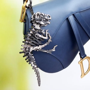 Puede incluir: Un bolso de hombro azul marino con forma de silla de montar, que presenta un dije de metal dorado en forma de D colgando de la correa frontal y un llavero de esqueleto de dinosaurio articulado en tonos negro y plata en el lateral.