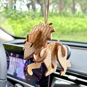 Könnte beinhalten: Ein hölzerner Löwen-Autoanhänger, der am Rückspiegel hängt. Der Löwe ist aufwendig mit einem Wabenmuster und braunen Kanten geschnitten. Das Innere des Autos ist sichtbar, mit Armaturenbrett und Lenkrad.