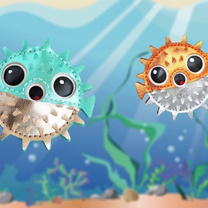 May include: Two puffer fish shaped accessories featuring large black eyes and small red mouths. The left fish is teal with a metallic gold lower half, while the right fish is orange with a metallic silver lower half, both covered in small spikes.