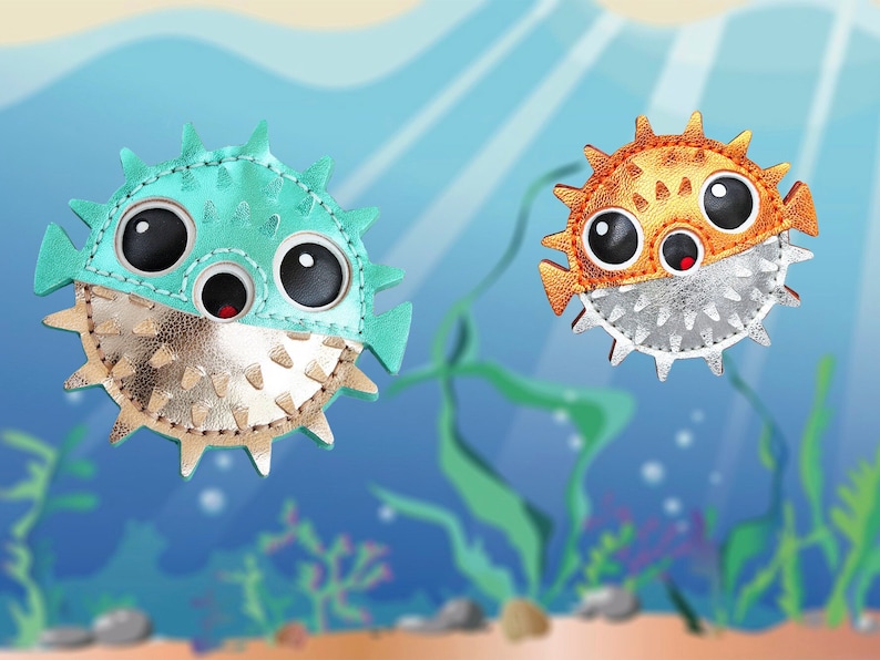 May include: Two puffer fish shaped accessories featuring large black eyes and small red mouths. The left fish is teal with a metallic gold lower half, while the right fish is orange with a metallic silver lower half, both covered in small spikes.