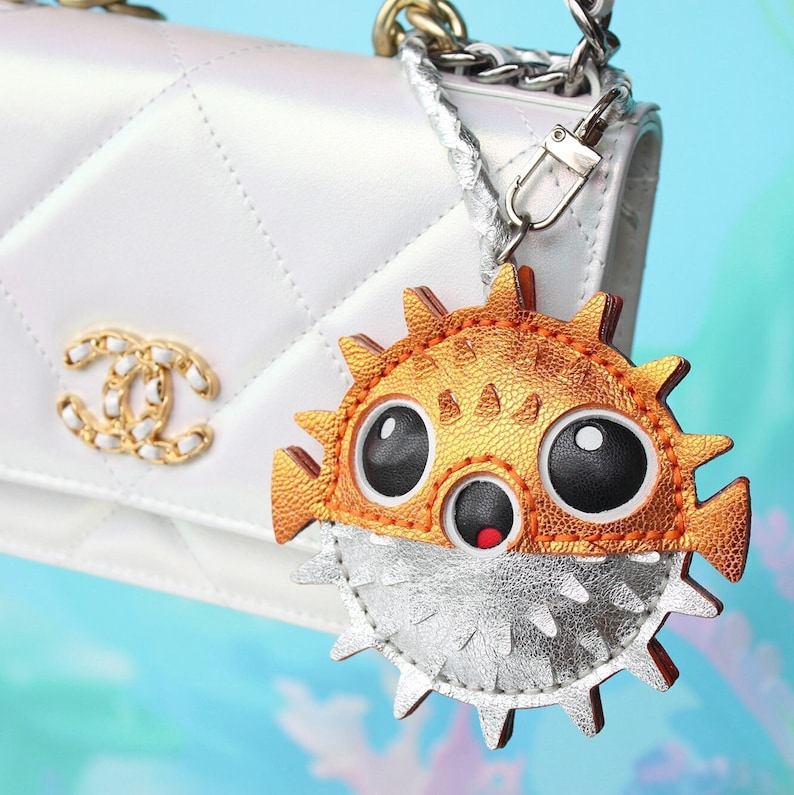 May include: A white quilted handbag with a gold-tone interlocking logo clasp and a silver chain strap. Attached to the strap is a pufferfish-shaped charm featuring a metallic orange upper section with large black eyes and a silver lower section.