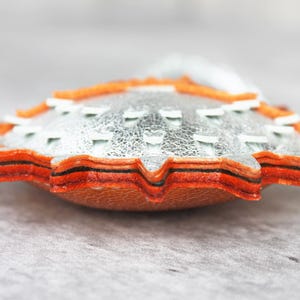 May include: A layered leather ornament featuring a metallic silver top layer with white stitched accents, stacked over multiple orange and brown leather layers with a serrated edge, creating a thick, textured, and multi-dimensional circular shape.