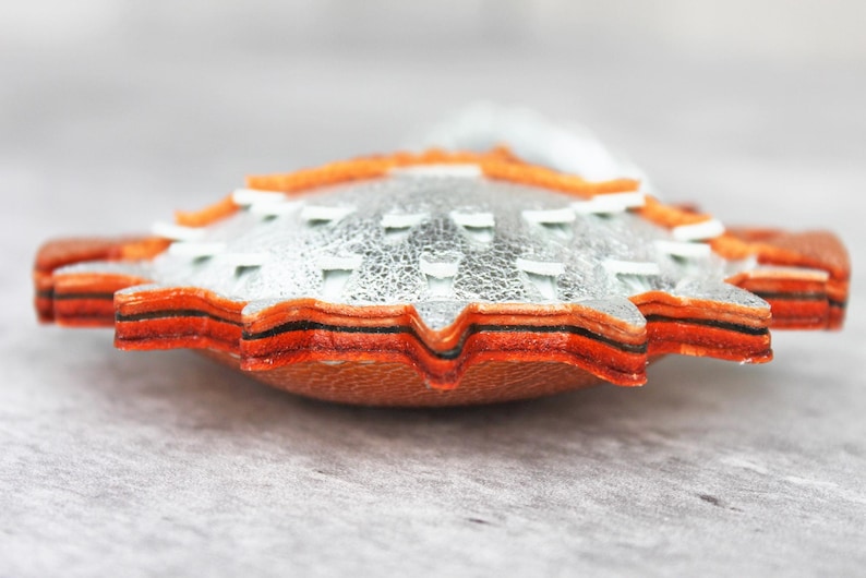 May include: A layered leather ornament featuring a metallic silver top layer with white stitched accents, stacked over multiple orange and brown leather layers with a serrated edge, creating a thick, textured, and multi-dimensional circular shape.