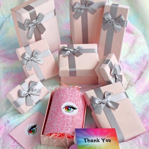 May include: A collection of light pink gift boxes wrapped with silver satin ribbons and bows. One open box contains pink leopard print tissue paper and shredded filler. Includes a thank you card and a heart-shaped card with a QR code.