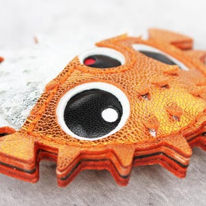 May include: A pufferfish-shaped keychain crafted from layered orange and silver textured material. The design features large, circular black eyes with white pupils and a jagged, spiky perimeter. A metal ring is attached to the side of the fish.